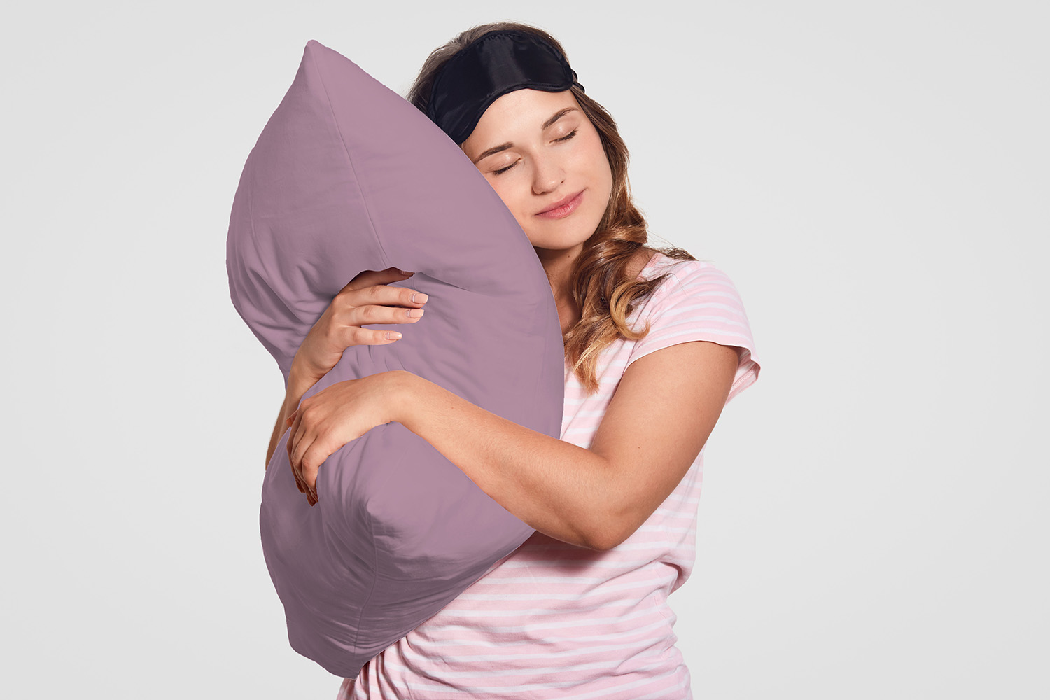 Photo of attractive European woman with healthy skin leans on soft pillow, wears pyjamas, eyewear on head, poses alone against white background, has sleepy look. People, good morning concept
